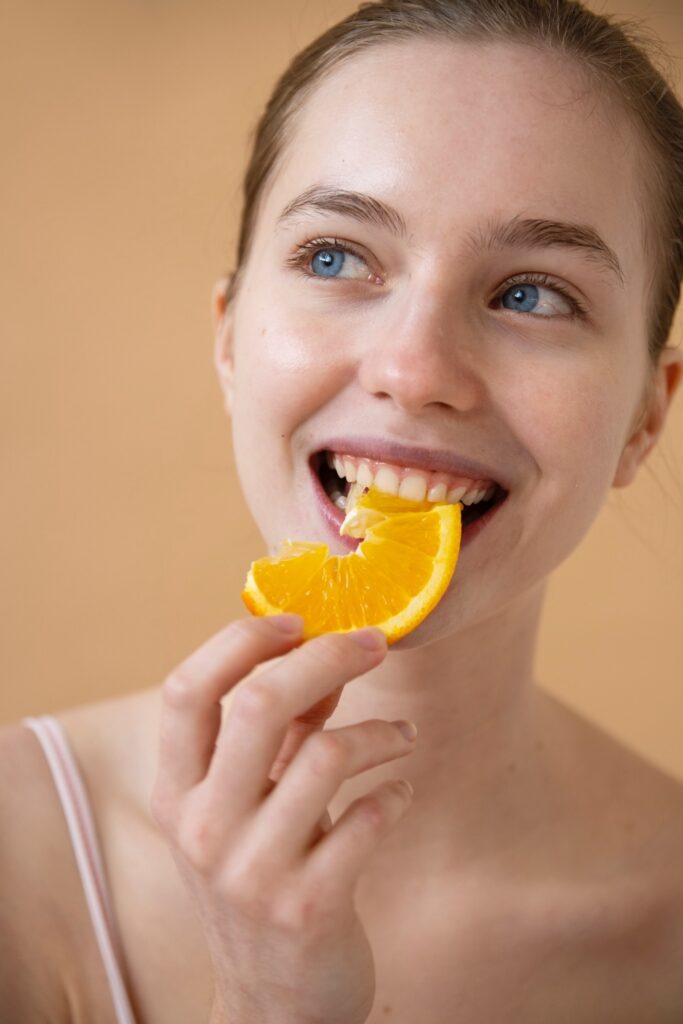 Vorderansicht einer lächelnden Frau mit einem Zahnfleischlächeln (Gummy Smile), die eine Orangenscheibe isst. Dies lässt sich mit Botox behandeln.