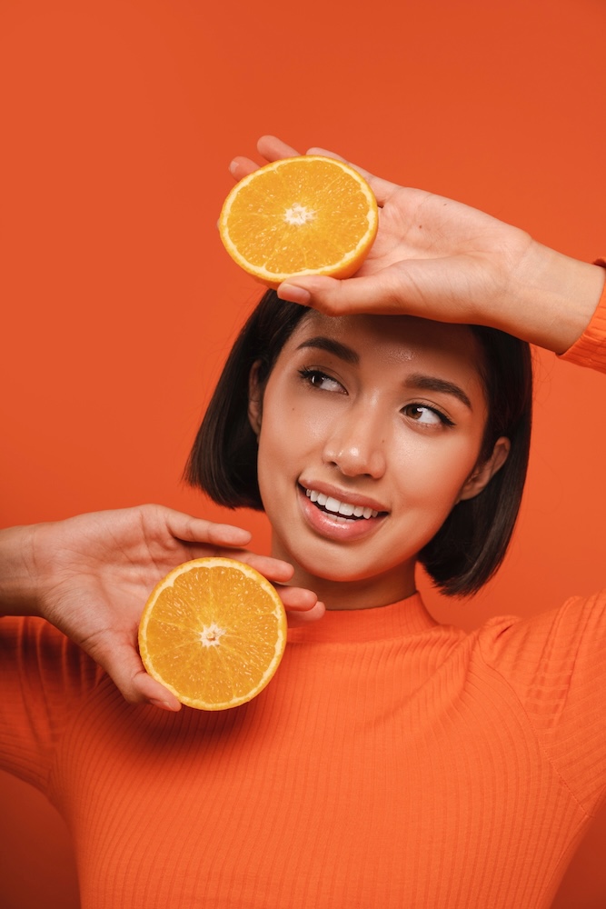Eine lächelnde Frau hält zwei Orangenscheiben vor einem kräftigen orangefarbenen Hintergrund. Das Bild symbolisiert Frische, Energie und die kraftvolle Wirkung von Vitamin C, wie sie in der Immune IV Drip Therapie enthalten ist.